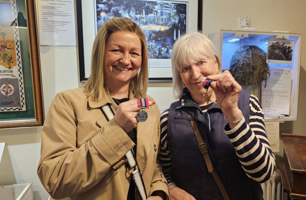 Colour photograph of two ladies holding war medals. On the right is Tracy Anderson and blonde woman in her 30s and on the left her mother Maggie
