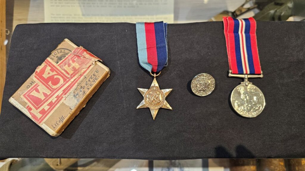 Colour image showing, from left to right, the buff cardboard box the medals arrived in a star shaped medal with white, red and blue ribbon, a button badge, a round medal with red, white and blue ribbon