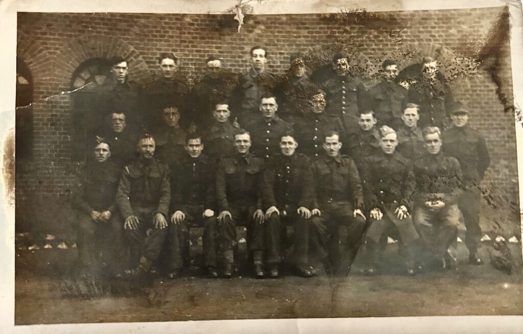 Black and white photograph of uniformed POWs. Three tows of men. The first row is seated. 24 men are in the photograph