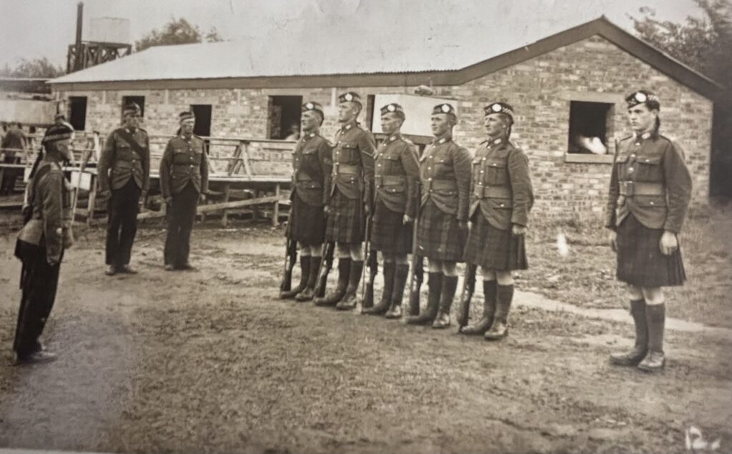black and white photograph of five soldiers on parade in a rural setting with one officer to the rear of the photograph. Soldiers are stood to attention and wearing Highland uniform with kilts