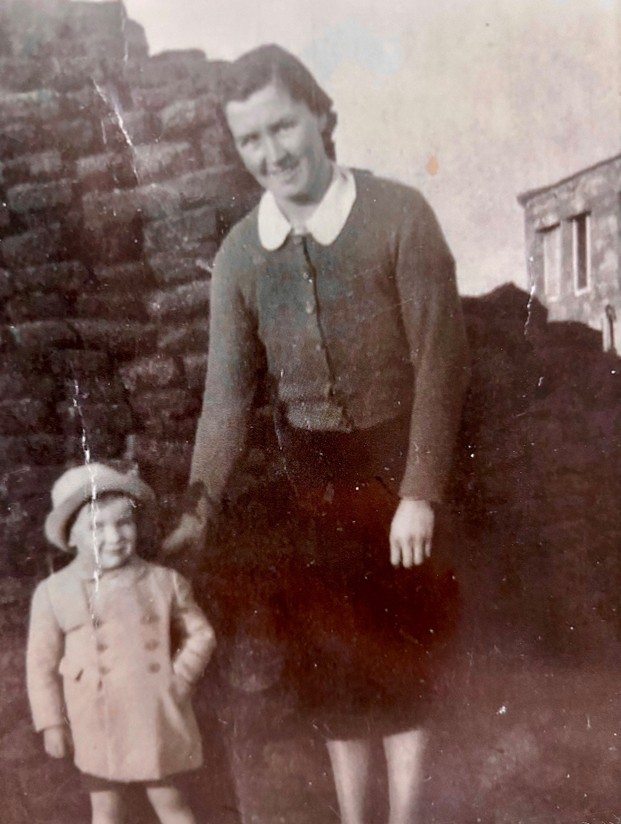 Black and white photograph of a young boy with his mother with a brick wall and house in the background. The photograph was taken in 1941 and the people are dressed in clothing appropriate for the period