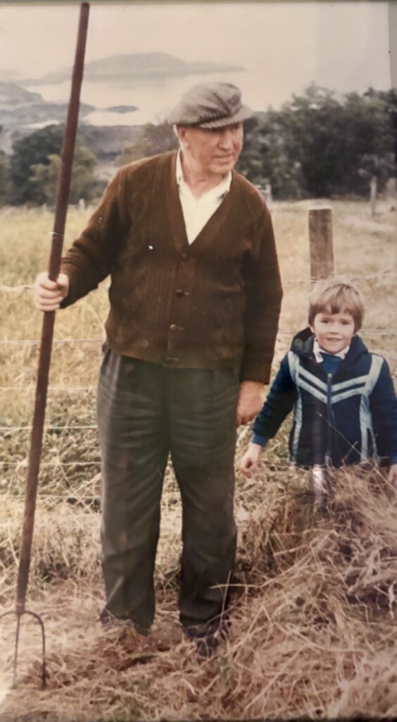 A colour photograph. An old man in a flat cap and brown cardigan stands with a young child of around five years old to his right. The boy has short brown hair and is wearing a blue anorak