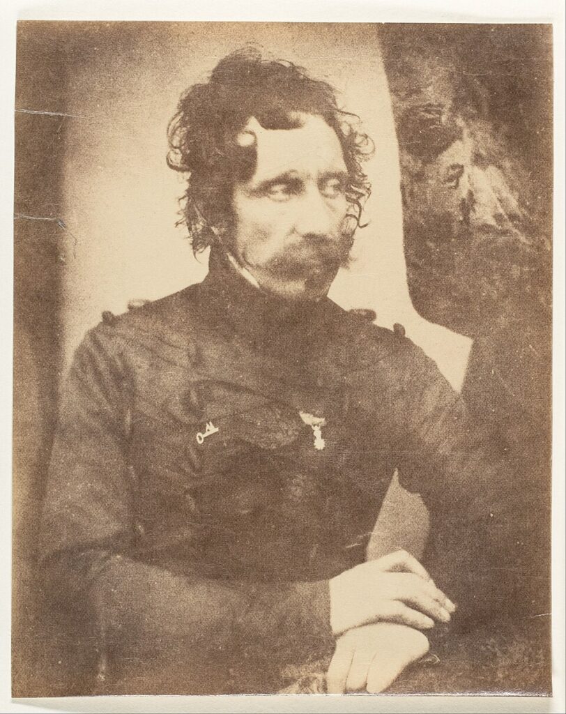 Sepia photograph of a man in Victorian military attire. He has curled hair and a handle bar moustache. The is looking to the right in the photograph