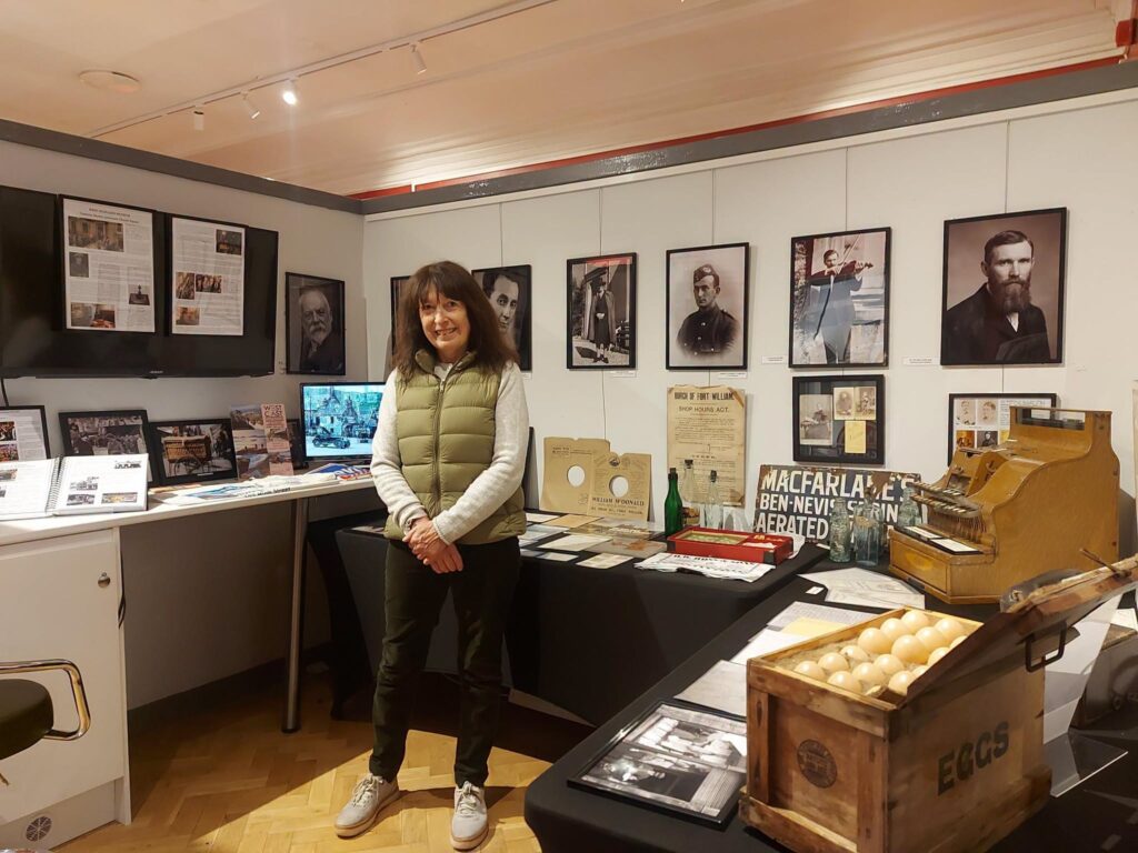 Colour photograph of a petite white women with dark shoulder length hair stood in front of a museum exhibition displaying framed photographs and objects such as a shop till and a TV showing a video